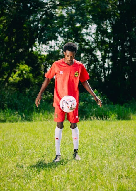 Male soccer player in red uniform practicing with a football on a green field outdoors.