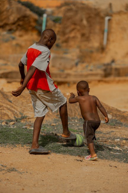 Two children play soccer on a dusty field, sharing joy and movement.